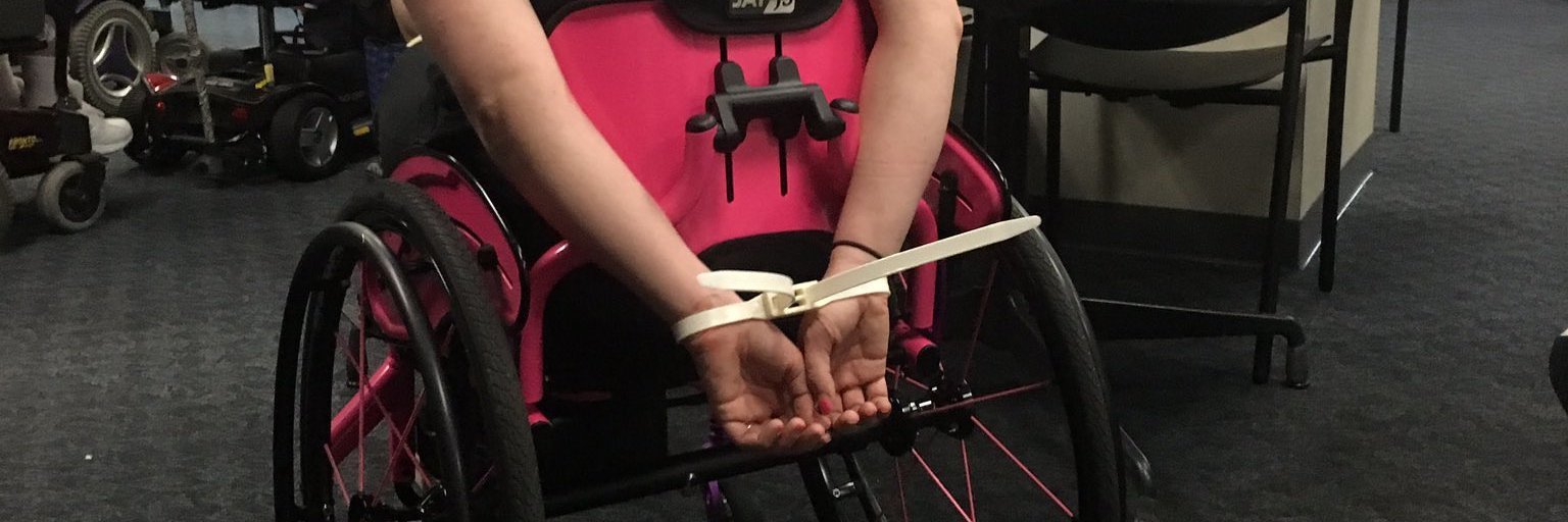 Disability rights activist Stephanie Woodward sits in her pink manual wheelchair with her hands cuffed behind her back.