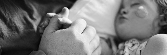 Black and white photo of little girl in hospital bed holding on to parent's hand while she sleeps