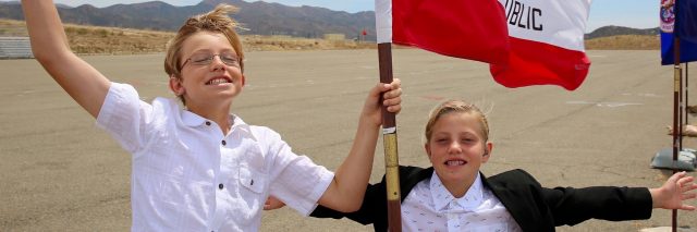 Two kids wearing white shirts and waving a flag.