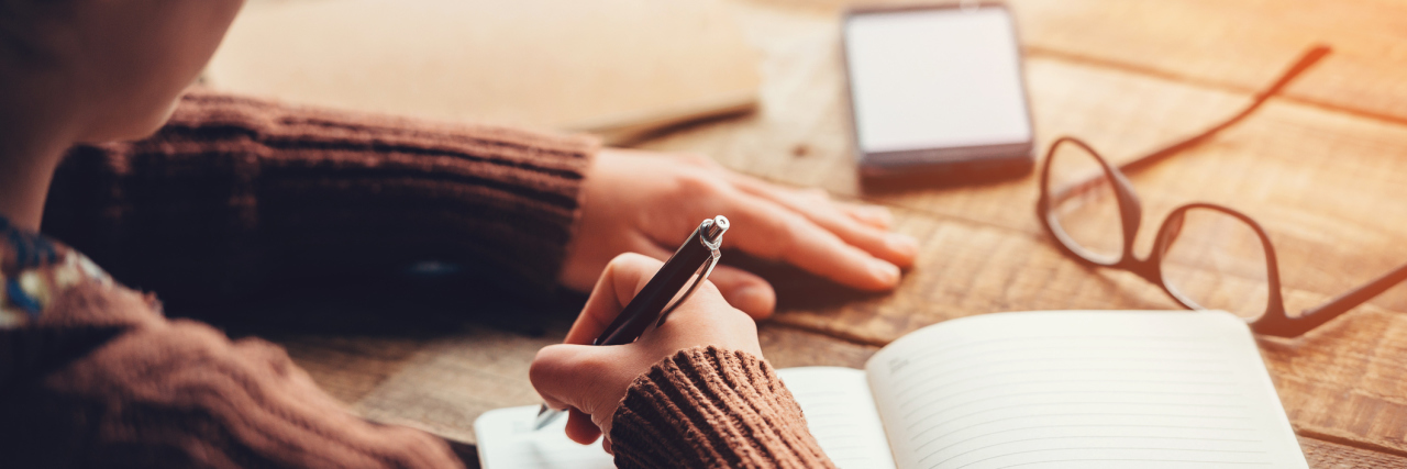 close up over shoulder shot of woman writing in notebook with glasses and phone visible on wooden table