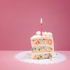 Slice of Colourful Birthday Confetti Cake with a lit candle over a pink background.