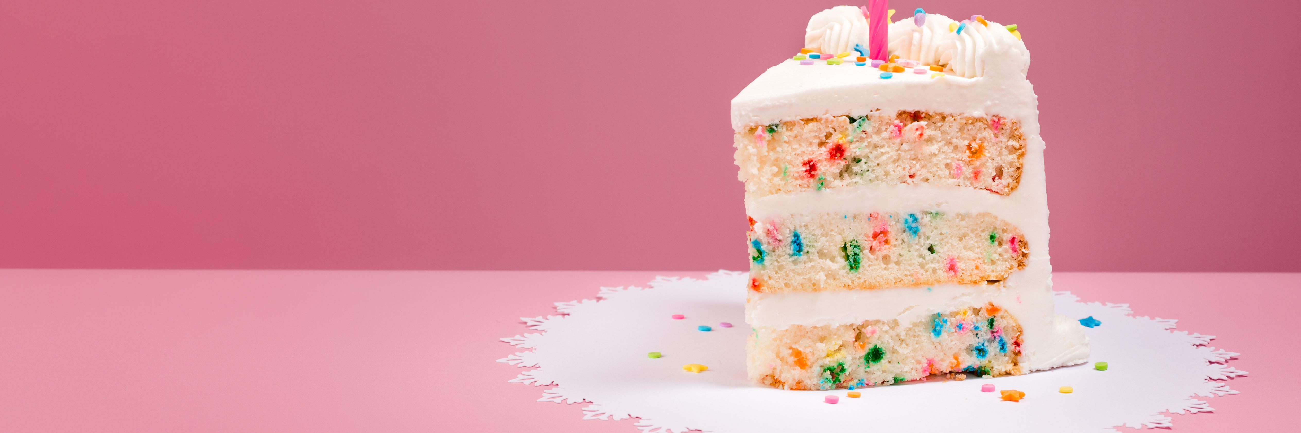 Slice of Colourful Birthday Confetti Cake with a lit candle over a pink background.