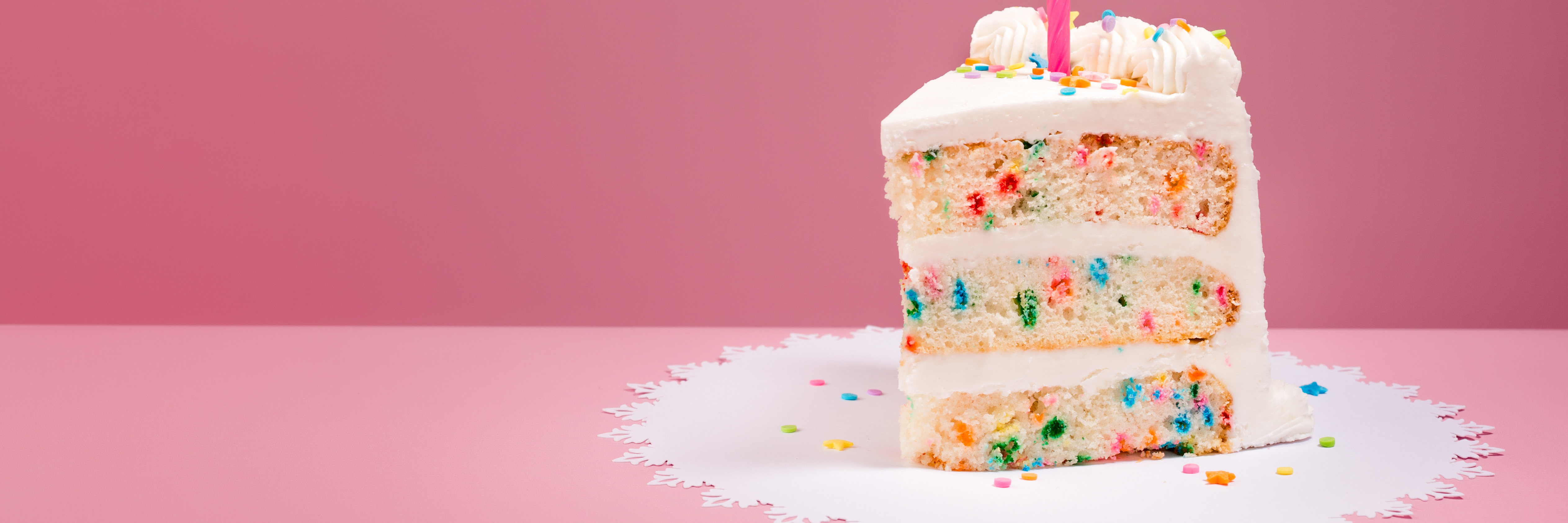 Why Birthdays Are Hard for Me as Someone With Mental Illness Slice of Colourful Birthday Confetti Cake with a lit candle over a pink background.