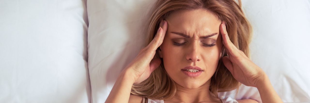 What I Wish My ER Doctor Knew About My Migraines A woman in bed, with her hands up to her head, grimacing in pain.