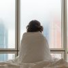 woman sitting on a bed wrapped in a blanket and looking out the window