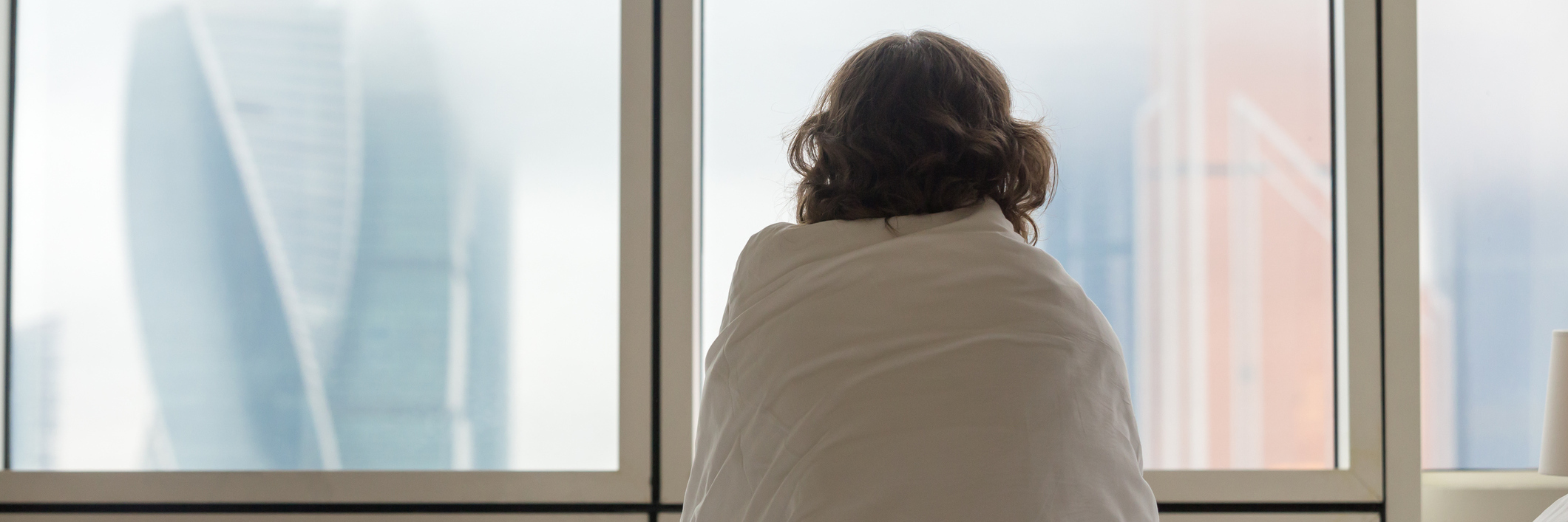 woman sitting on a bed wrapped in a blanket and looking out the window