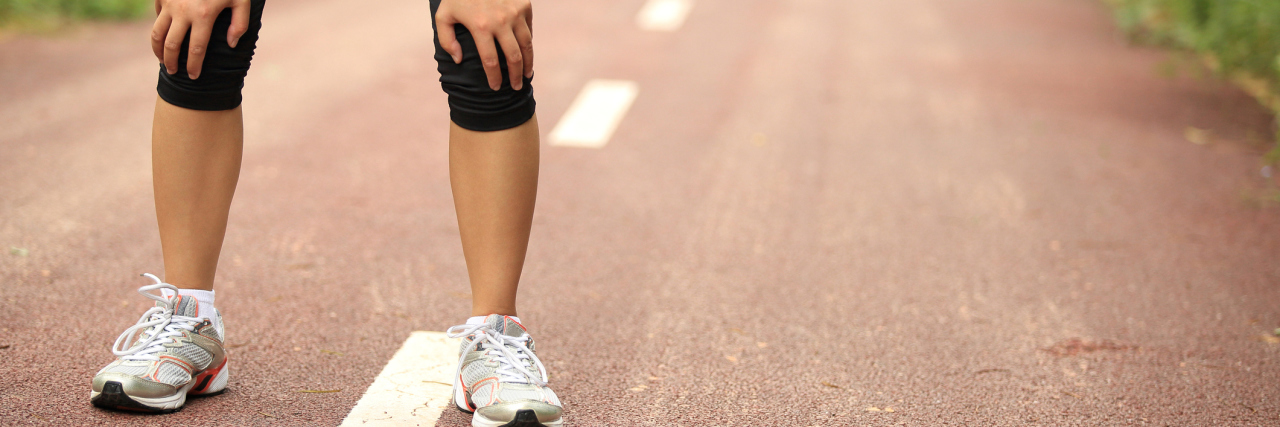 Why I Tell People Chronic Illness Is 'a Marathon, Not a Sprint' A tired female runner, taking a break from her run with her hands on her knees.