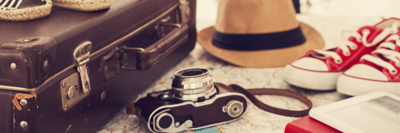 A map in the background with a suitcase, hat, camera, and other travel gear on top.