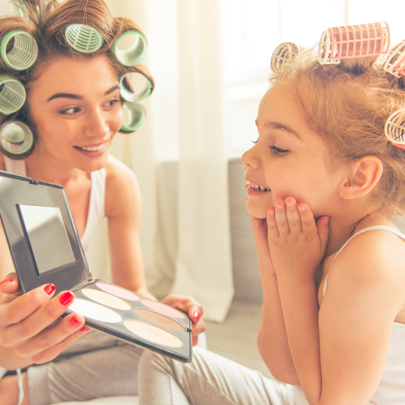A mom and her young daughter, wearing curlers, looking into a mirror.