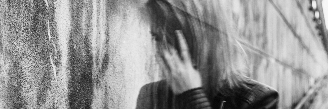 Young woman suffering from a severe disorientation, confusion, or sadness outdoors, in front of a wall. Converted to black and white, grain added, blurry, slightly out of focus.