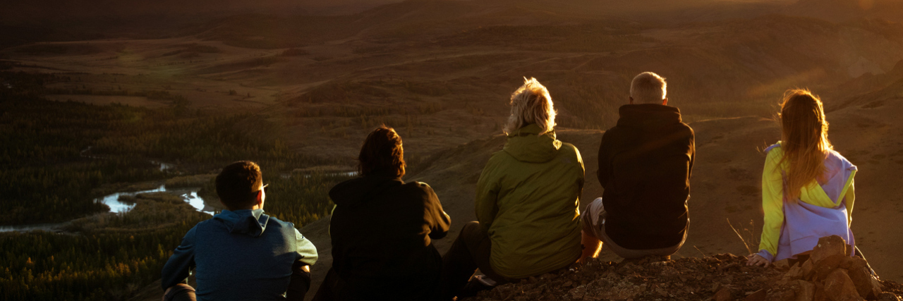 Group of friends sits on the mountain's top on background of mountains and sunset