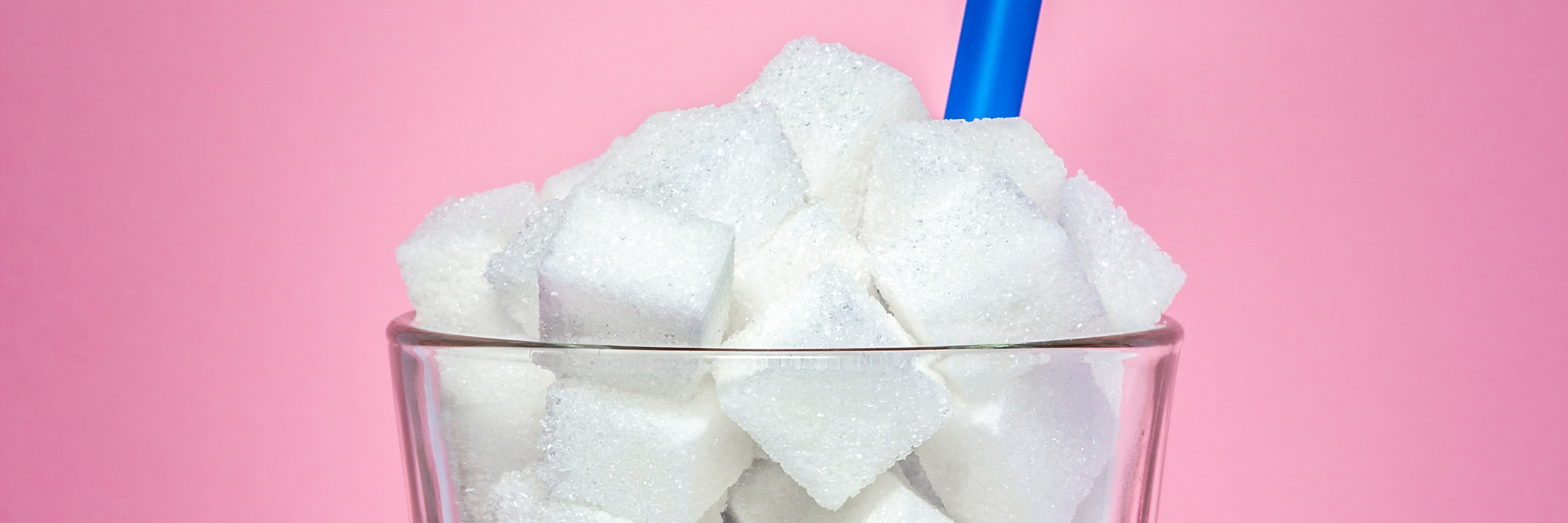 What You Need to Know About the Study Linking Sugar to Depression Glass full of sugar cubes on a pink background with a blue straw