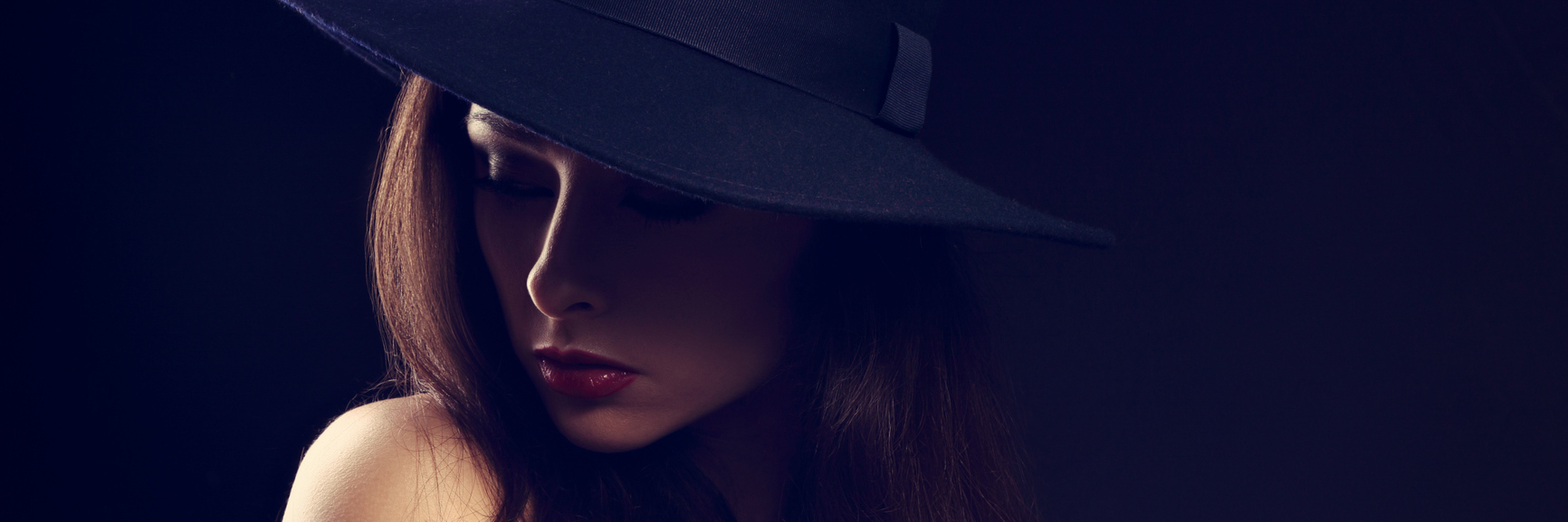 A woman wearing a hat, looking down, while surrounded by darkness.