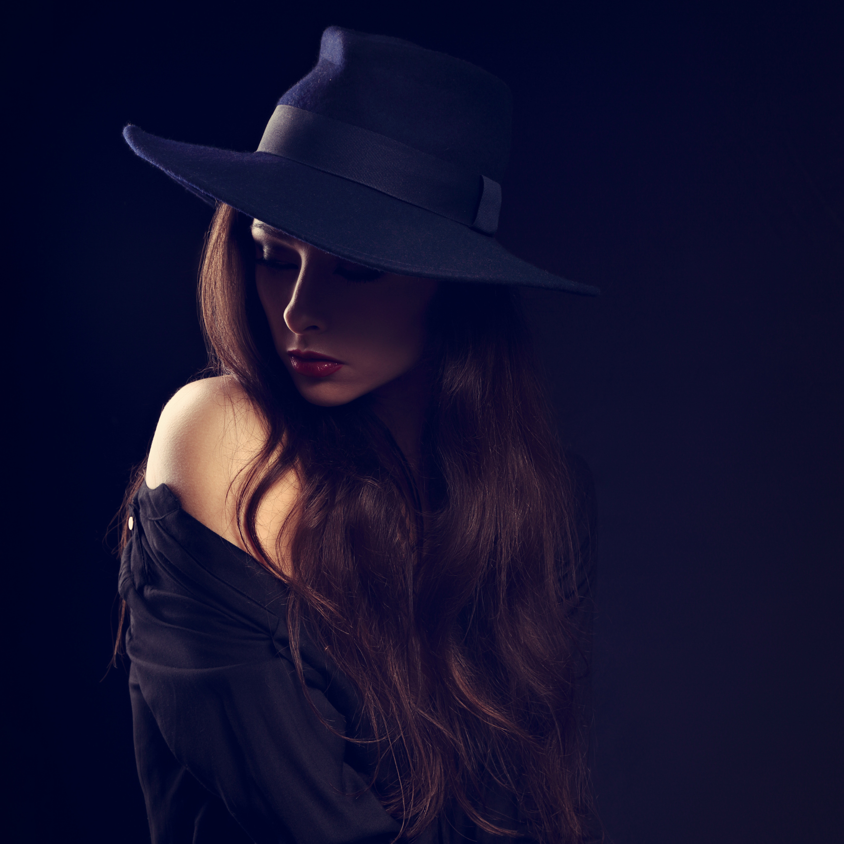 A woman wearing a hat, looking down, while surrounded by darkness.
