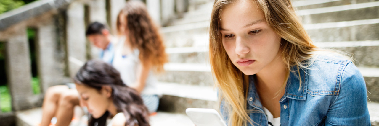 4 Tips to Help Make Life at School Easier If You Have Celiac Disease A teenage girl sitting on steps, looking at her phone, with other young students blurred behind her.