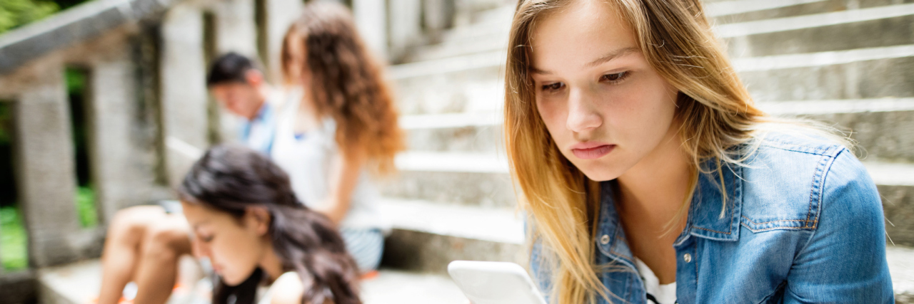 A teenage girl sitting on steps, looking at her phone, with other young students blurred behind her.