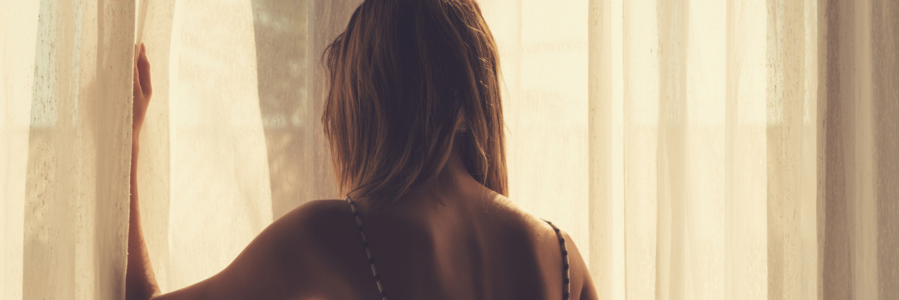 The Part of My Job That Affected Me as Someone in Self-Harm Recovery sepia tone photo of girl holding curtain open in room with back turned to camera