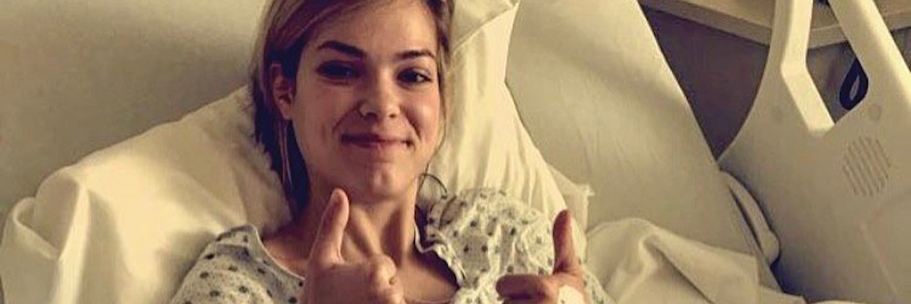 The writer of the article laying in a hospital bed, giving two thumbs up.