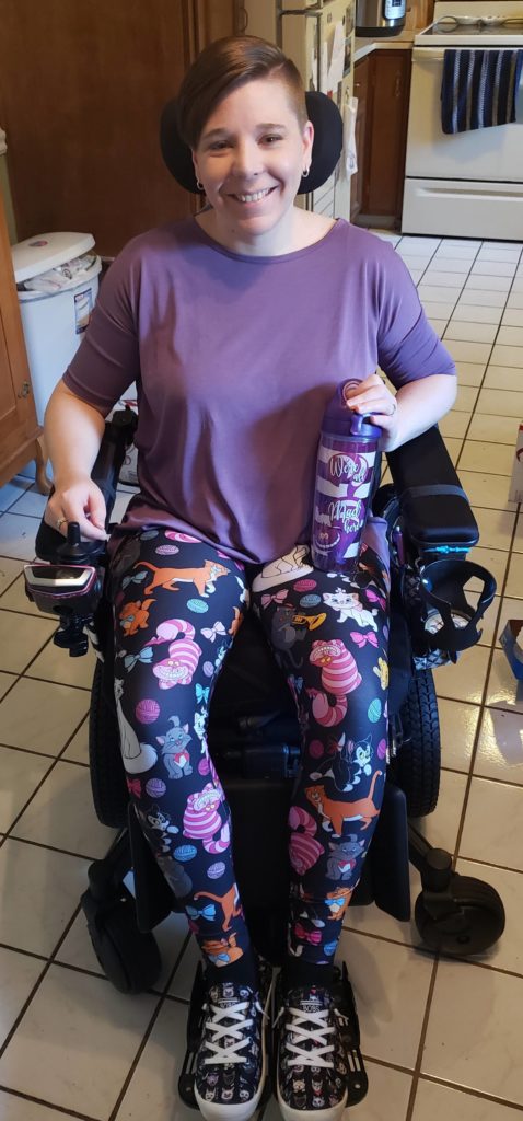 Me sitting in my wheelchair in my kitchen wearing purple leggings picturing all Disney cats, purple shirt, and purple Cheshire Cat Tervis water mug.
