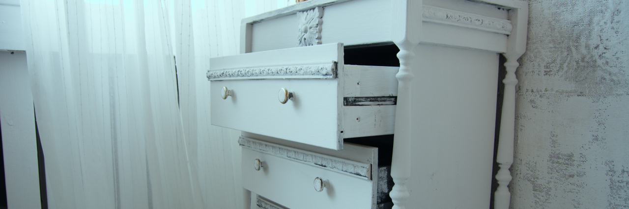 An old painted white ladies chest of drawers with open drawers by the window. Ladies' retro table near the cloth wall