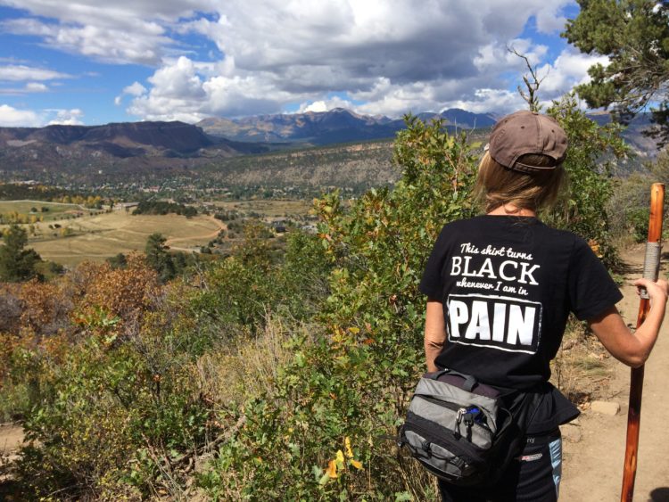 woman hiking in the mountains with a black shirt on that says 'this shirt turns black whenever I am in pain'