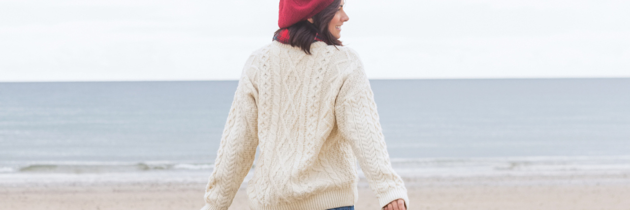 Why I'm Going to Stop Apologizing for My Epilepsy rear view of woman wearing hat and sweater, walking on beach on a cloudy day