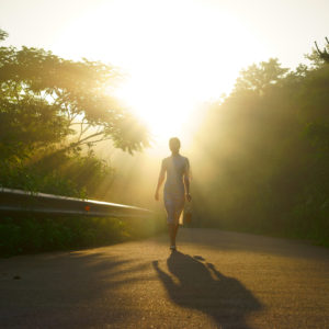 woman walking down a trail between trees with sunlight ahead