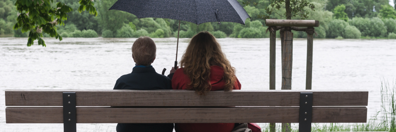 How to Support People With Chronic Illness Who Have Suicidal Thoughts two women sitting on a park bench next to a lake under an umbrella