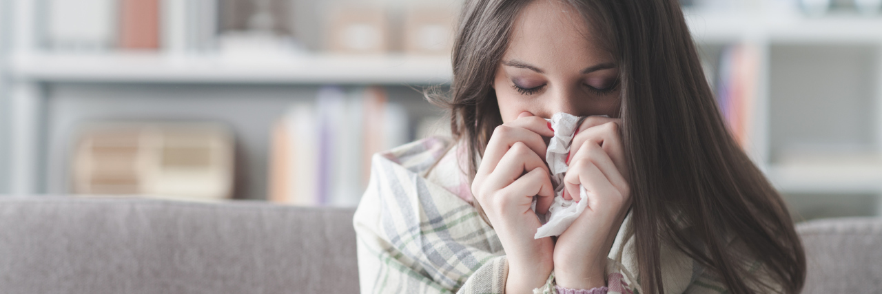 Why Cold and Flu Are Harder to Fight When You're Chronically Ill Sick young woman at home on the sofa with a cold, she is covering with a blanket and blowing her nose