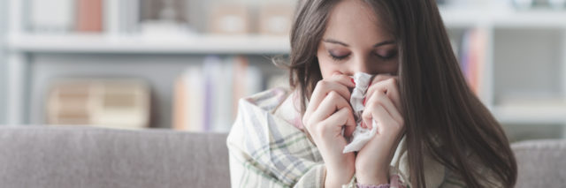 Sick young woman at home on the sofa with a cold, she is covering with a blanket and blowing her nose