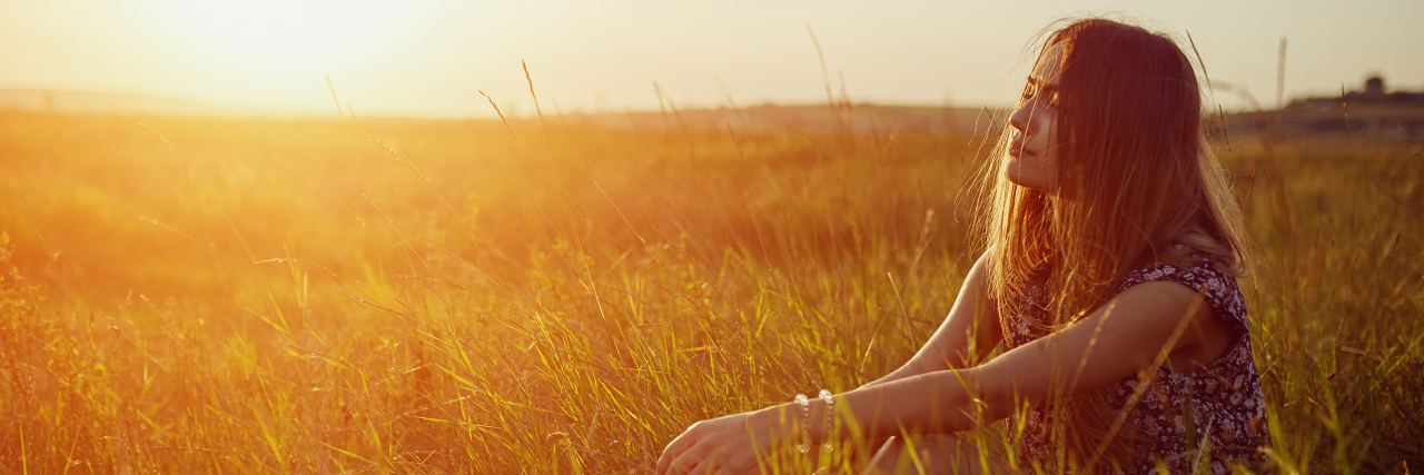 Being a Caregiver for Someone With a Chronic Illness A woman sitting alone in a field at sunset, surrounded by tall grass and warm golden light.