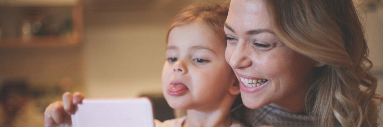 A mom and daughter taking a selfie together, the mom smiling and child sticking her tongue out.