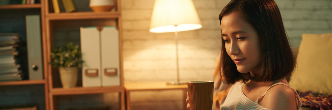 Why I Have a Chronic Illness Hibernation Bubble An Asian woman sitting on a couch, reading a book and drinking a hot drink.