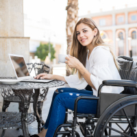 A young woman at a cafe, writing on her laptop, as she sits in her wheelchair.