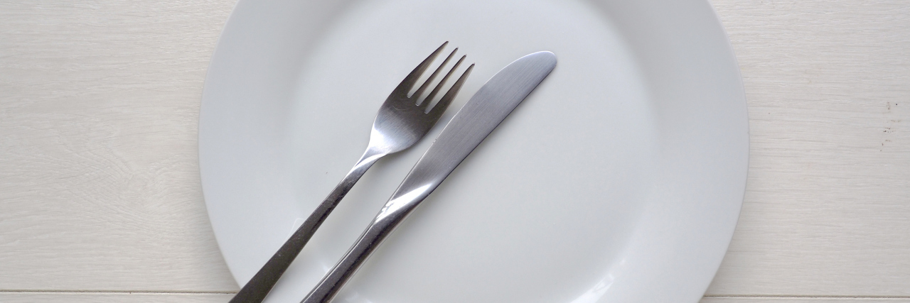 These Diet Trends Aren't Revolutionary, They Glorify Anorexic Behavior White plate and knife and fork on white wooden background