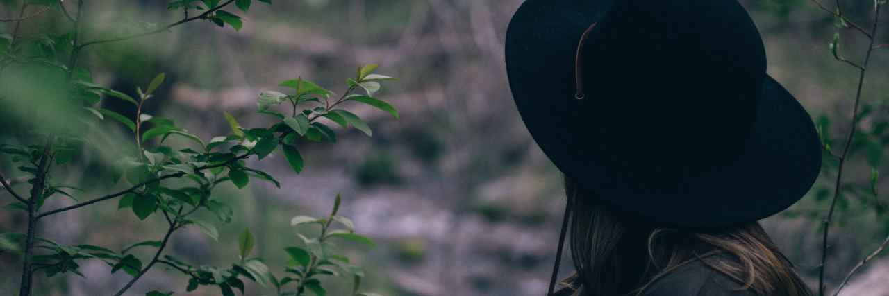 woman in large black hat stands in the woods looking away from the camera