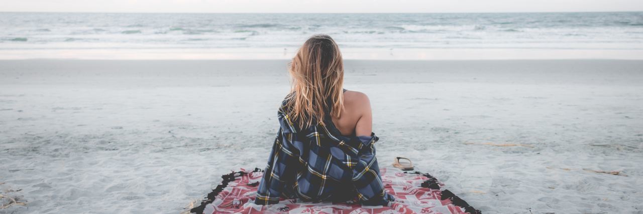 The Importance of Patience During Health Struggles With Migraine woman sitting on a blanket on the beach looking at the ocean