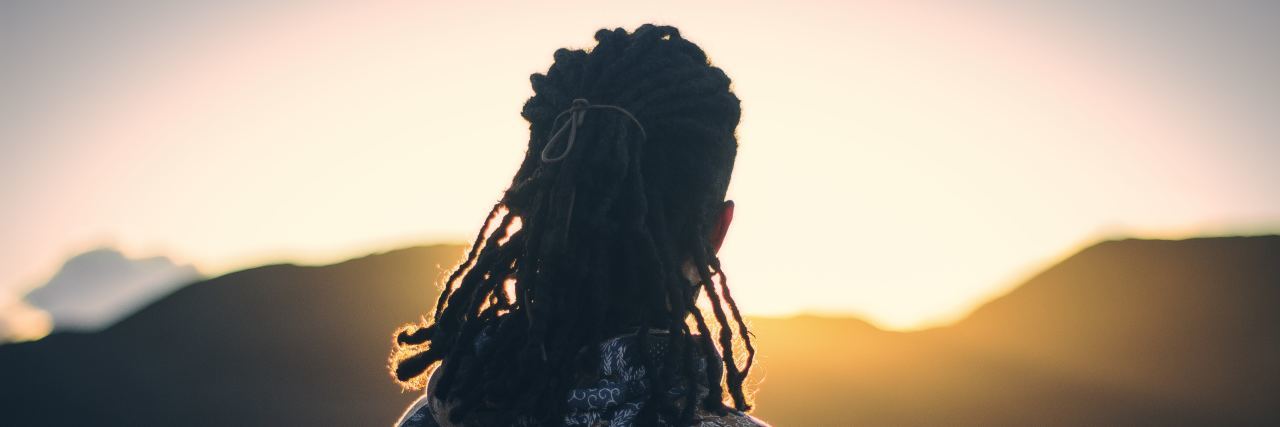 a man with dreadlocks stands on a mountain with a sun setting