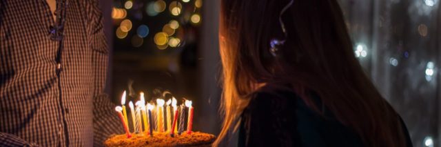 man holds birthday cake with lit candles as woman blows the candles out