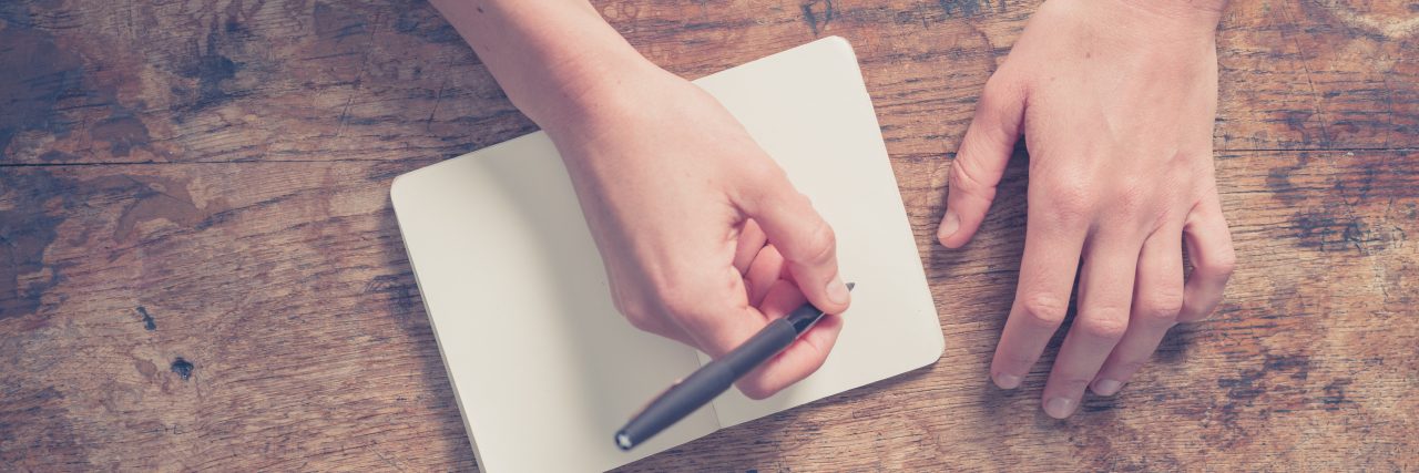 Letter to Boy With Shwartz Jampel Syndrome Close up on the hands of a young woman as she is writing in a small notepad at a wooden table