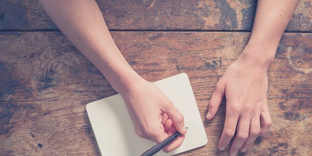 Close up on the hands of a young woman as she is writing in a small notepad at a wooden table