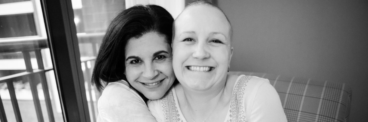 Two female friends, one with a bald head, hugging.