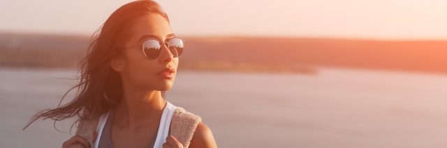 A woman wearing sunglasses, with a sunset and body of water behind her.