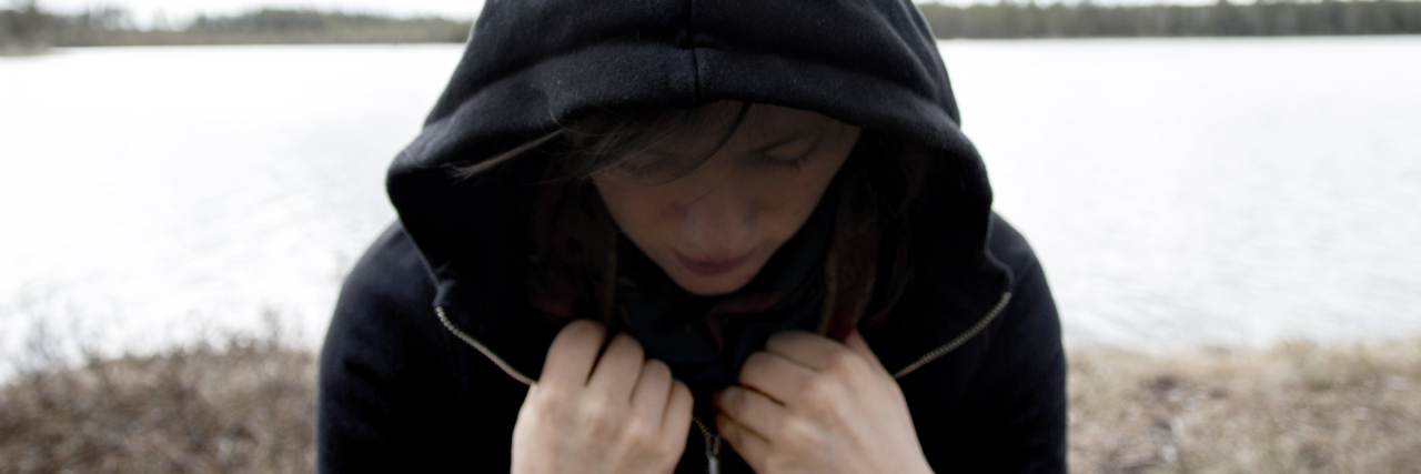 Woman in a black hoodie standing with the back towards a lake. She is looking down and has a shadow over her face. Concept photo of depression, mental illness and loneliness.