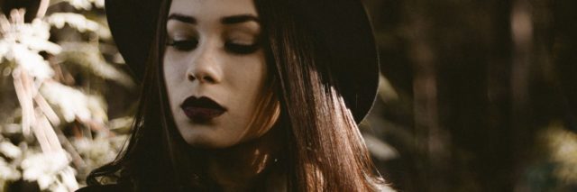 woman with dark hat and dark lipstick stands in a forest