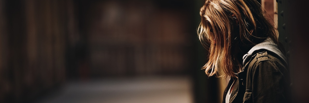 30 Things People Said That Were Actually Code for 'I'm Depressed' A depressed woman leans against wall in dimly lit hallway, looking down with her hair obscuring her face.