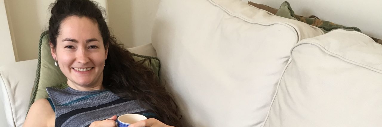 How to Make Working With Illness Easier The writer laying on her couch with a cup of tea in her hand, and her cat sitting on her lap.