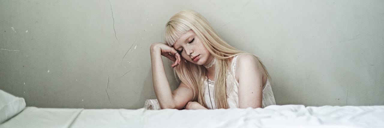 blonde woman sitting beside bed depressed against white wall
