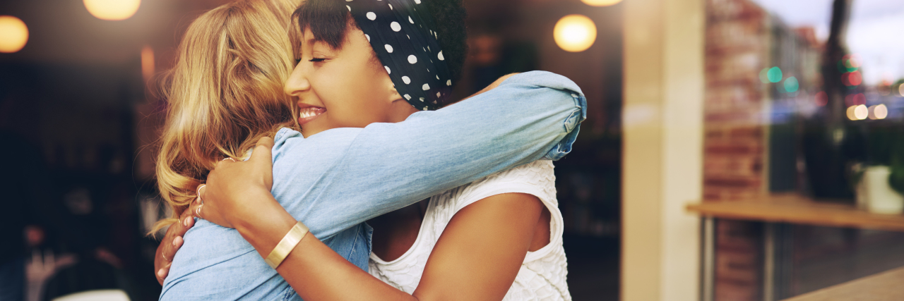 Two multi ethnic affectionate girl friends embracing as they sit in a coffee shop enjoying a cup of coffee together