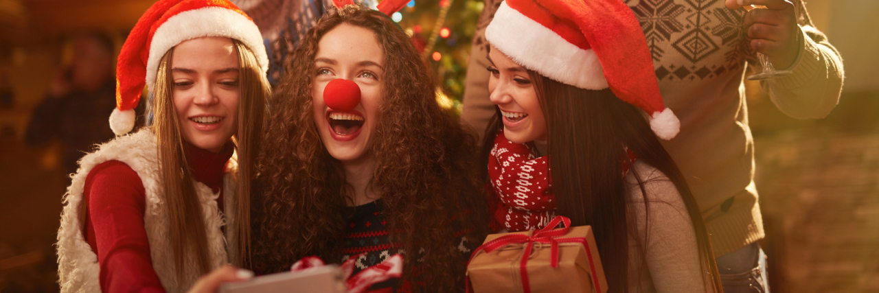 How I Afford the Christmas Season While Living With a Chronic Illness A group of friends taking a selfie in front of the Christmas tree, holding gifts and wearing antler headbands and Santa hats.